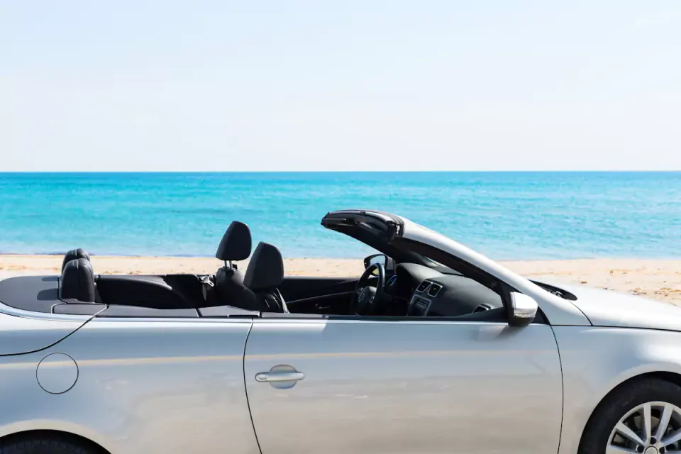 Carro cabrio prateado com a capota aberta, estacionado junto à praia e ao mar